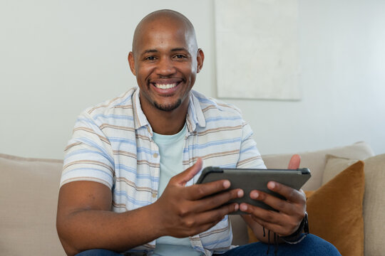 African man sitting on beige sofa in living room holding tablet and smiling at camera