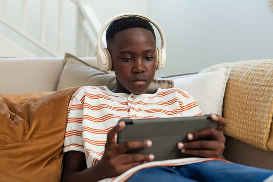 African teenage male sitting on sofa in living room, wearing white headphones and using tablet