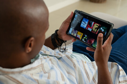 Adult African man reclining on sofa at home, holding tablet in case, in striped shirt, bracelets