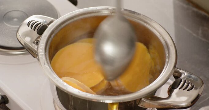 Spoon entering frame is stirring round orange slices in stainless pot on stove, preventing sticking