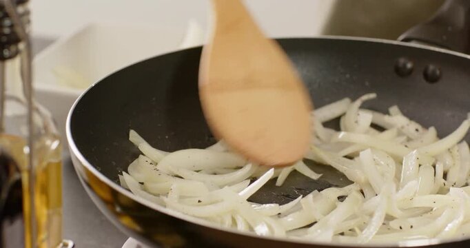 Wooden spatula entering frame is stirring sliced white onions in nonstick pan to soften and glaze
