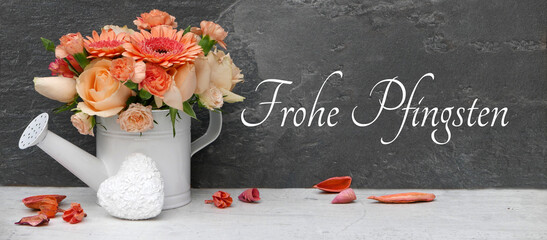 Gießkanne mit Rosen und Gerbera, davor ein weißes Herz und verstreute Blütenblätter vor dunklem Hintergrund mit dem Schriftzug 'Frohe Pfingsten'