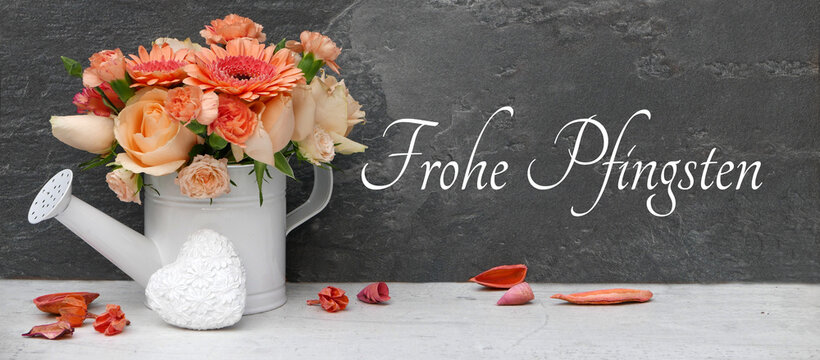 Gie&szlig;kanne mit Rosen und Gerbera, davor ein wei&szlig;es Herz und verstreute Bl&uuml;tenbl&auml;tter vor dunklem Hintergrund mit dem Schriftzug 'Frohe Pfingsten'
