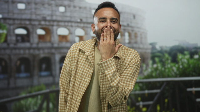Man in front of coliseum building blows a kiss with hand to mouth while smiling at camera; friendly greeting joy.