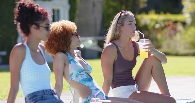 Female friends chatting on pool deck, sipping cup, noticing off-camera movement, center in swimsuit