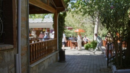 Fototapeta premium Soft bokeh outdoor cafe scene with shaded terrace, umbrella tables, stone walkway and leafy trees, shallow defocused foreground and background; background backdrop copyspace backplate calm.