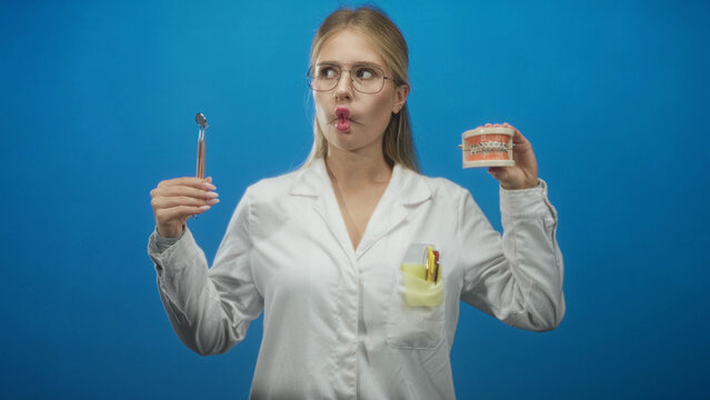 Woman dentist in white coat holds dental mirror and orthodontic braces model while puckering lips fish face gesture in studio; playful confidence.