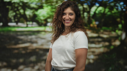 Fototapeta premium Woman smiling, wearing white t shirt with bare forearms visible, standing in a forest among green trees and dappled light; happiness friendly approachable.