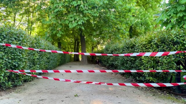 Park pathway blocked with red and white warning tape, restricted access in green garden alley, safety barrier outdoors