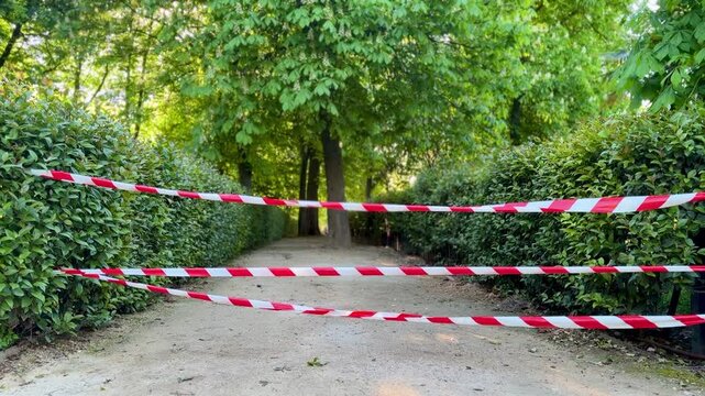 Park pathway blocked with red and white warning tape, restricted access in green garden alley, safety barrier outdoors