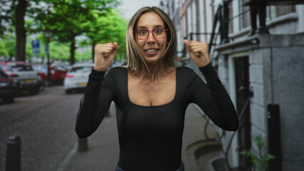 Obraz premium Woman wearing red glasses, smiling with fists raised and visible cleavage on a busy city street; victory celebration.