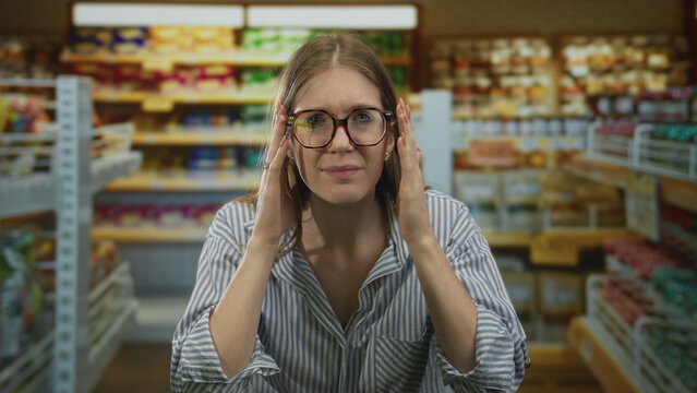 Blonde woman adjusts her glasses with both hands between colorful shelves in brightly lit supermarket aisle; concentration.