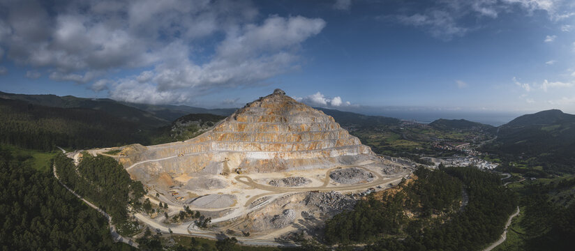 Aerial view of the massive terraced open-pit quarry surrounded by lush green forests and rolling hills under a cloudy blue sky in Santa Lucia de Gordon, Castile and Leon, Spain.
