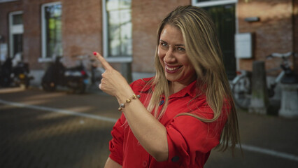 Fototapeta premium Woman in red blouse smiling broadly with finger pointing into frame on street; optimism energy engagement joy.