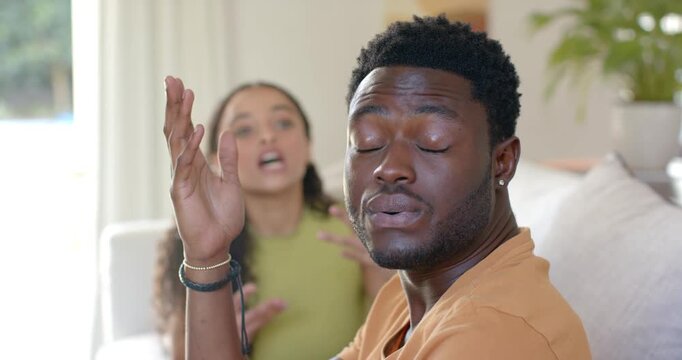 African American couple on couch woman speaking behind, man in tan tee raising hand, rubbing temple