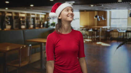 Obraz premium Woman wearing santa hat smiling and looking up inside a building cafe with wooden tables and chairs; cheerful holiday.