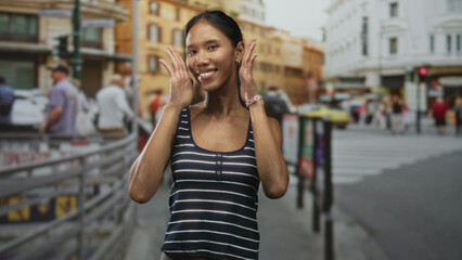 Fototapeta premium Thai woman waving hands and smiling with face visible on a busy city street near buildings and crosswalk, wearing striped tanktop; travel joy.