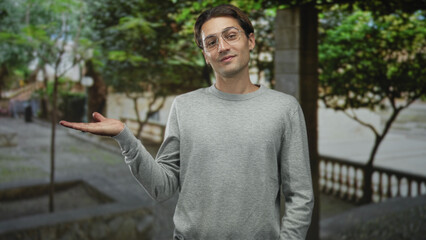 Fototapeta premium Man presenting palm up with open hand and wrist gesture, wearing glasses and grey sweater on a street; welcoming ease.