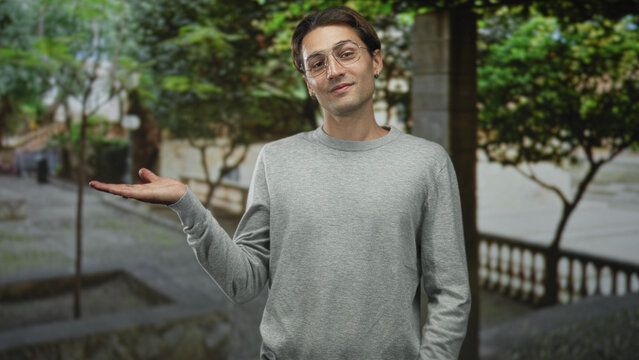 Man presenting palm up with open hand and wrist gesture, wearing glasses and grey sweater on a street; welcoming ease.