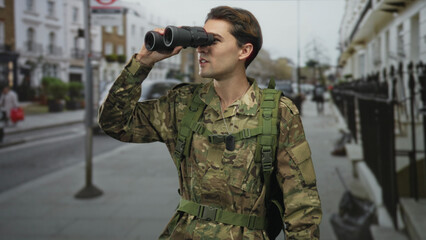 Soldier man in camouflage holds binoculars to eyes on a street, dogtags and backpack visible;...