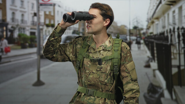 Soldier man in camouflage holds binoculars to eyes on a street, dogtags and backpack visible; vigilant duty.