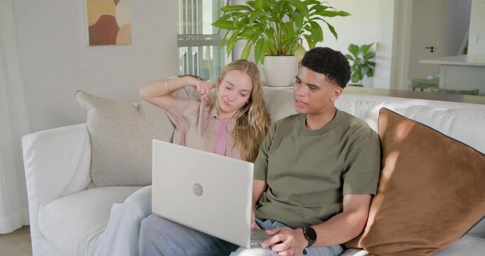 Diverse couple sitting on sofa at home, woman pointing at silver laptop, man typing to respond
