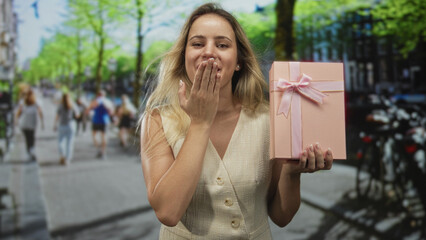 Fototapeta premium Young blonde woman holding pink gift box with ribbon, blowing a kiss with hand to lips on a busy city street with blurred pedestrians and bicycles; giving celebration joy.