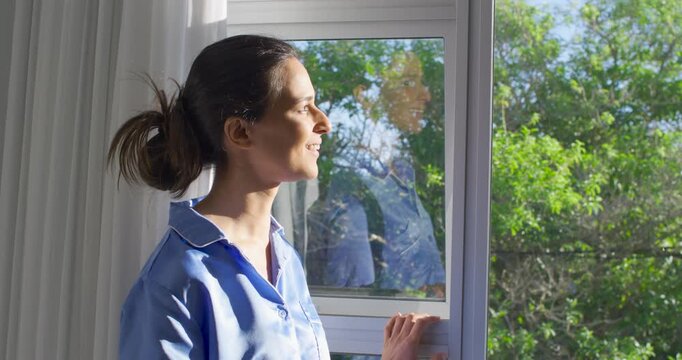Adult female standing at home window holding sash facing sun feeling heat reflecting white curtains