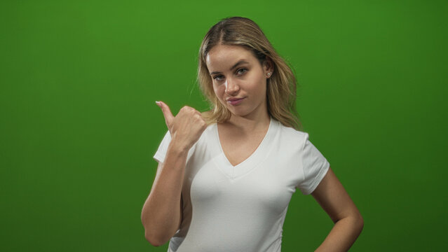 Woman young blonde in white t shirt pointing thumb over shoulder in green studio, smirking with hand on hip; playful confidence.