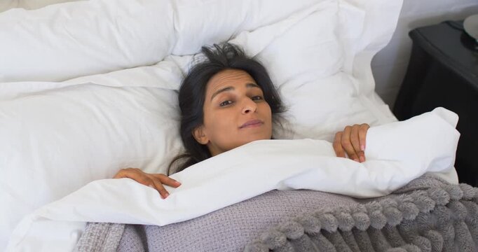 Indian woman waking from sleep, opening eyes, peeking over duvet, smiling and laughing in bed