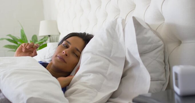Indian adult woman wearing blue sleepwear waking in bed to white alarm clock ringing and silencing