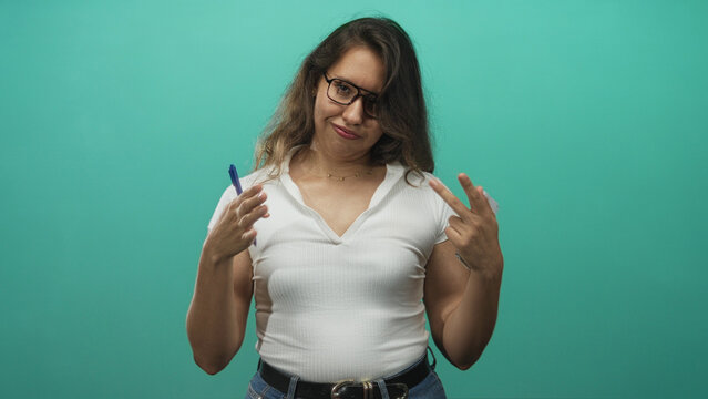 Young hispanic woman in white shirt points fingers to chest while holding pen and notepad in teal studio; sarcasm attitude.
