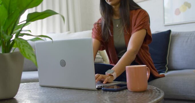 Woman typing on laptop on sofa, reaching for pink mug, sipping during break while glancing phone