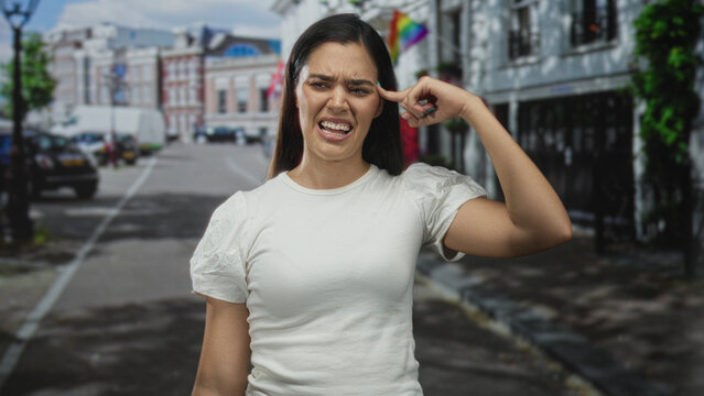 Woman points finger to temple on city street while grimacing at noise and traffic; frustration disbelief.