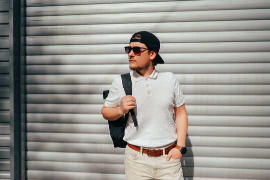Young White Man Wearing Sunglasses And Cap, Leaning Against Metal Shutter, Backpack Over Shoulder, Holding