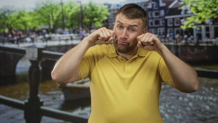 Fototapeta premium Young overweight man in yellow polo pinches cheeks and pulls a silly face on a street canal bridge in amsterdam by a boat; playful.