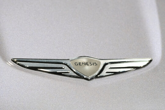 Genesis car emblem badge on a silver metallic hood, chrome winged logo centered on a smooth painted surface, luxury automotive branding, macro detail of texture and reflective finish