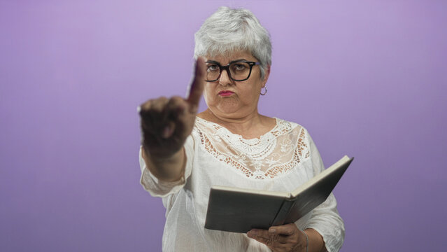 Woman reader with glasses holds an open book and extends her hand palm forward in a studio purple set as a stop gesture; determination.