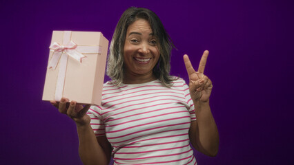 Obraz premium Middleaged hispanic woman holding a pink gift box, flashing a peace sign with her hands and smiling in studio; celebration joy.