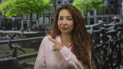 Fototapeta premium Woman pointing finger on street beside a canal in amsterdam with parked bicycles and building facades visible, frowning expression; doubt.