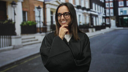 Fototapeta premium Woman judge in black robe wearing glasses, smiling with hand to chin and crossed arm pose on a quiet street lined with townhouses; thoughtful confidence.