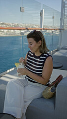 Obraz premium Woman sipping cocktail on cruise terrace, hand holding glass with pineapple garnish and sunglasses on head while seated on sofa; summer getaway relaxation.