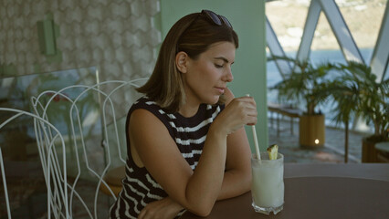 Obraz premium Woman stirs drink with straw at a table in a building by the water, sunglasses on head; contemplation moment.