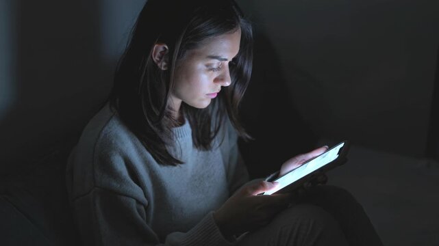 Jeune femme seule dans une pi&egrave;ce sombre &eacute;clair&eacute;e par la lumi&egrave;re bleue du smartphone, expression fatigu&eacute;e et d&eacute;filement lent, ambiance cin&eacute;matographique minimaliste &eacute;voquant l&rsquo;addiction num&eacute;rique