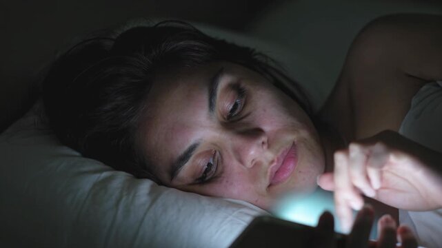 Gros plan sur une femme allong&eacute;e dans son lit &eacute;clair&eacute;e par la lumi&egrave;re du smartphone, expression fatigu&eacute;e et d&eacute;filement lent, ambiance nocturne minimaliste &eacute;voquant insomnie et isolement