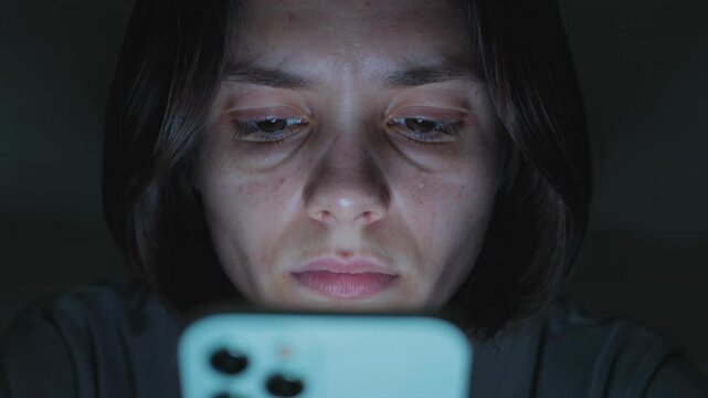 Gros plan sur un visage &eacute;clair&eacute; par la lumi&egrave;re bleue d&rsquo;un smartphone dans l&rsquo;obscurit&eacute;, mouvements subtils des yeux et expression fatigu&eacute;e, ambiance minimaliste et introspective