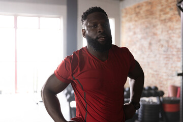 African American man pausing in gym near dumbbell rack and plates, in red Tshirt, wristwatch © wavebreak3