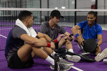 Three male athletes sitting on purple court near net, checking smartphone and holding padel rackets © wavebreak3