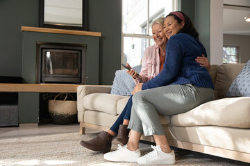 Naklejka premium Senior female partners sitting on living room sofa at home holding remote and smiling together