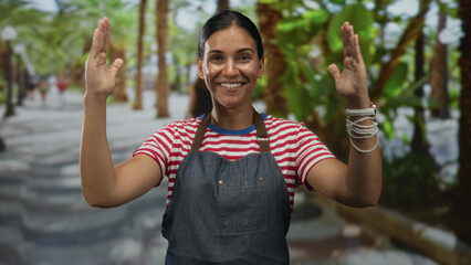 Naklejka premium Woman in denim apron and red striped shirt smiles and raises both hands palms up while standing on a tree lined street with palms; joy.